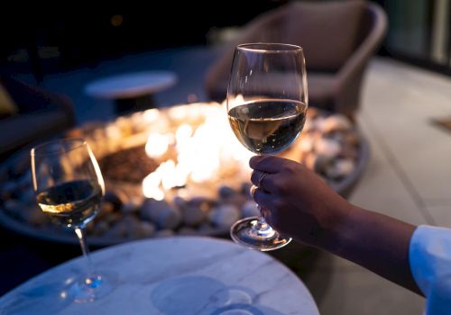 The image shows a hand holding a glass of wine near a fire pit, with another glass on a table in the foreground.