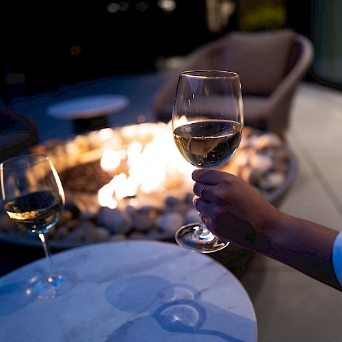 The image shows a hand holding a glass of wine near a fire pit, with another glass on a table in the foreground.