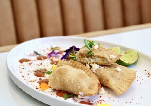 Three empanadas on a plate garnished with cheese, herbs, and lime; accompanied by colorful sauce drizzles and edible flowers.