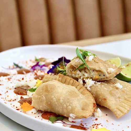 Three empanadas on a plate garnished with cheese, herbs, and lime; accompanied by colorful sauce drizzles and edible flowers.