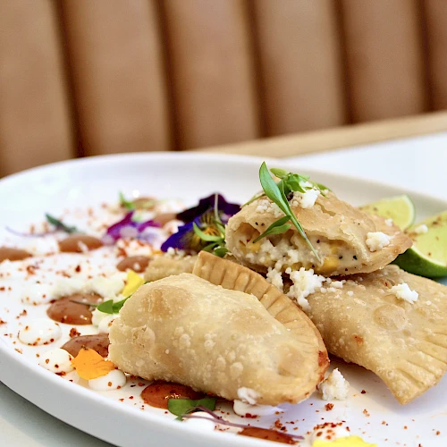 Three empanadas on a plate garnished with cheese, herbs, and lime; accompanied by colorful sauce drizzles and edible flowers.