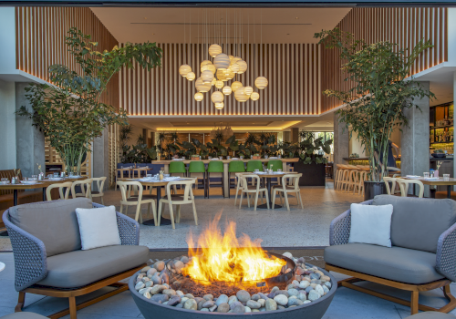 A modern restaurant with outdoor seating, a fire pit, plants, and a stylish interior with pendant lights and wooden furniture.