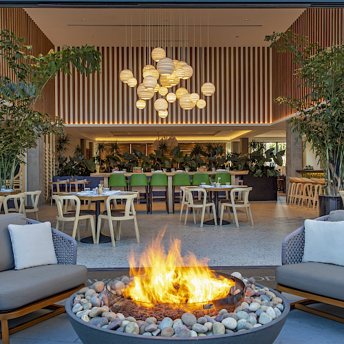 A modern restaurant with outdoor seating, a fire pit, plants, and a stylish interior with pendant lights and wooden furniture.