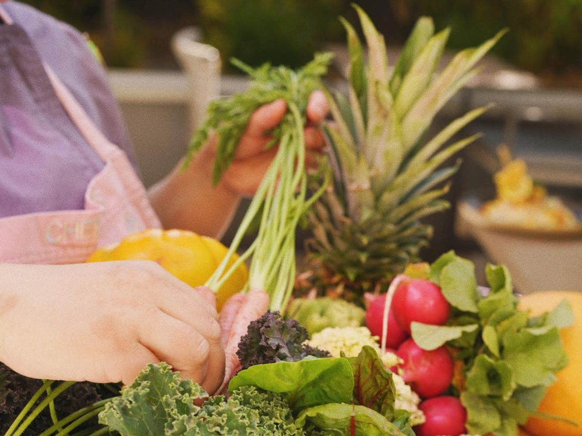 A person arranging a colorful assortment of fresh vegetables and fruits, including kale, peppers, radishes, and a pineapple, in a wooden bowl.