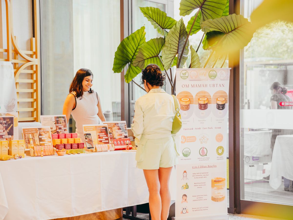 In a shop booth, two women browse OM Mamah ubtan skincare products beside jars, scrubs, and bright green leaves decor.