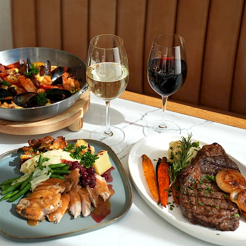 The image shows a paella, a plate with chicken and vegetables, another with steak and vegetables, plus glasses of white and red wine.