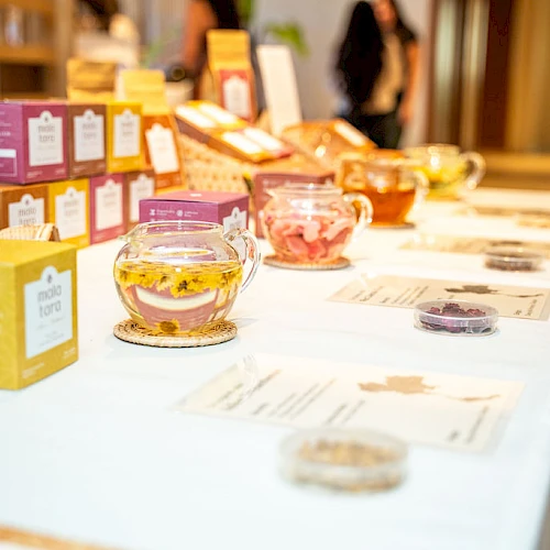A table with colorful tea packaging, glass teapots filled with loose tea, and informational cards arranged in a bright setting.