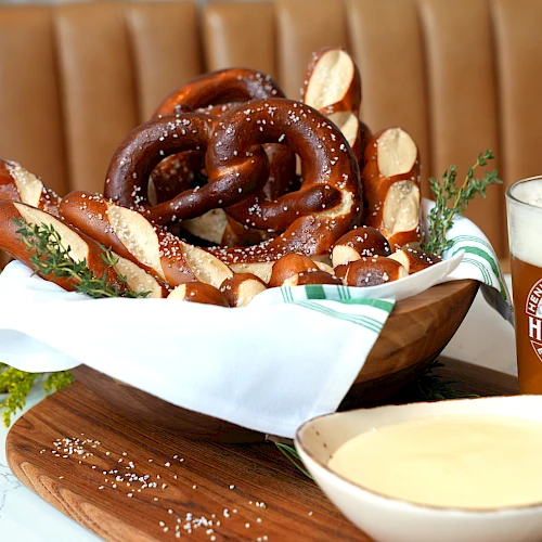 A basket of pretzels, a bowl of cheese dip, and a glass of beer are on a table with a brown booth background.