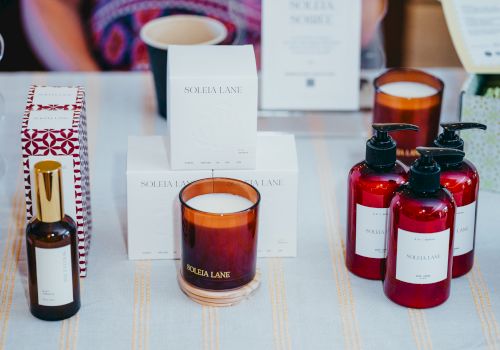 The image shows a display of scented candles, lotion bottles, and a spray bottle arranged on a table, with packaging in the background.