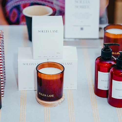 The image shows a display of scented candles, lotion bottles, and a spray bottle arranged on a table, with packaging in the background.