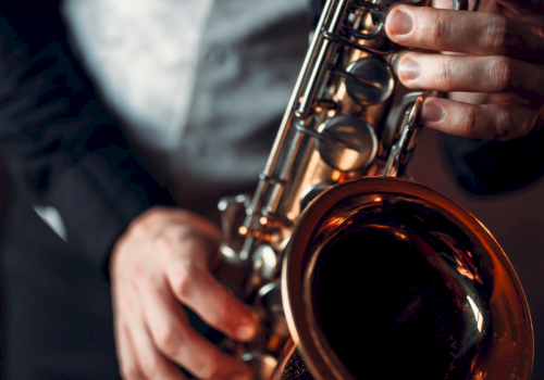 A person is playing a saxophone, wearing a vest and suit. The setting is dimly lit, possibly in a bar or lounge environment.