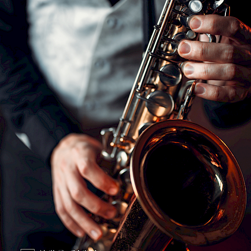 A person is playing a saxophone, wearing a vest and suit. The setting is dimly lit, possibly in a bar or lounge environment.