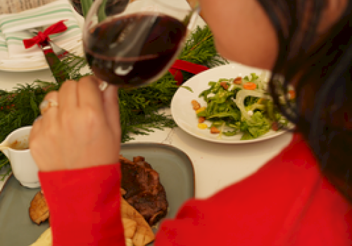 A person in red enjoys a glass of red wine at a festive dinner table with various dishes and a green candle lit.