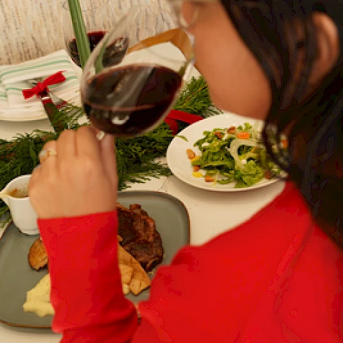 A person in red enjoys a glass of red wine at a festive dinner table with various dishes and a green candle lit.