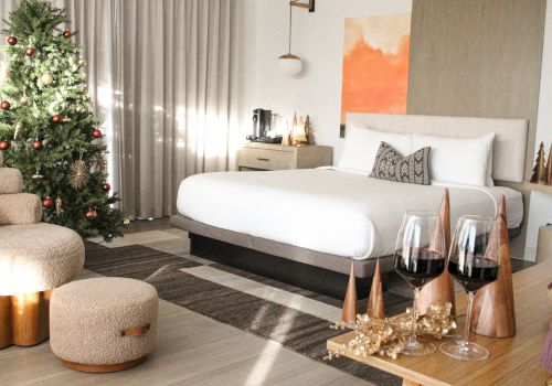 A cozy, modern bedroom with a Christmas tree, soft lighting, a large bed, a wooden side table, and festive decorations including pine cones and candles, ending with a chic vibe.