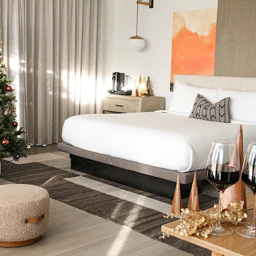 A cozy, modern bedroom with a Christmas tree, soft lighting, a large bed, a wooden side table, and festive decorations including pine cones and candles, ending with a chic vibe.