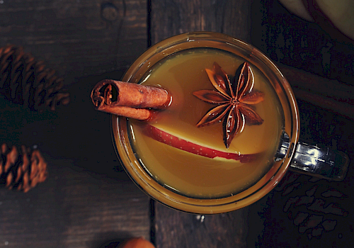 A warm beverage with cinnamon sticks and star anise, served in a mug on a dark wooden table with pine cones nearby.