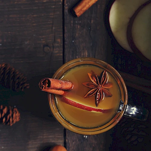 A warm beverage with cinnamon sticks and star anise, served in a mug on a dark wooden table with pine cones nearby.