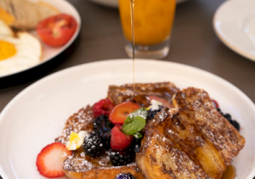 Pouring syrup over French toast with berries and mint on a plated breakfast dish, while a juice and toast sit in the background.