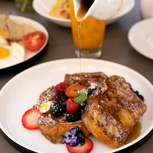 Pouring syrup over French toast with berries and mint on a plated breakfast dish, while a juice and toast sit in the background.