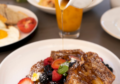 Golden French toast topped with berries; syrup streams over as cream pours, breakfast perfection on a sunny plate.