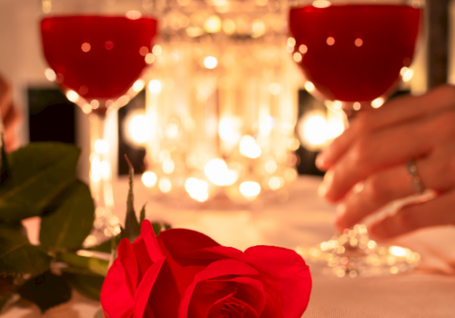 Two wine glasses, a red rose on a candlelit table, hands nearby, soft warm glow in the background.