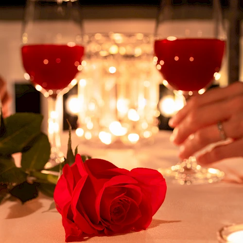 Two wine glasses, a red rose on a candlelit table, hands nearby, soft warm glow in the background.