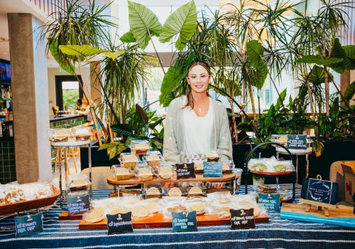 A friendly vendor stands behind a colorful market table with breads and cheeses, surrounded by lush indoor plants and signs labeling items.