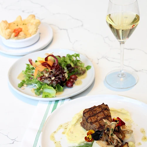 A gourmet plate with a seared steak over creamy sauce and mushrooms, a fresh greens salad, and a side of roasted potatoes, plus a glass of white wine.