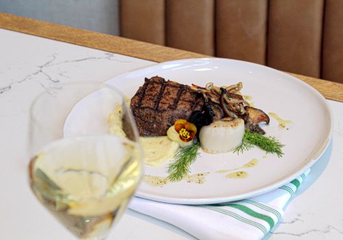 A plated steak with herbs and root vegetables, served with a sauce drizzle, alongside a glass of white wine, on a marble table.