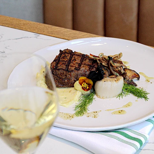 A plated steak with herbs and root vegetables, served with a sauce drizzle, alongside a glass of white wine, on a marble table.