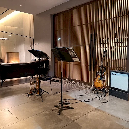 A cozy lounge with a music corner: microphones, stands, a guitar, amp, and sheet music on a stand in front of wooden slats and soft seating area.