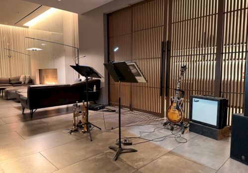 A cozy lounge with a music corner: microphones, stands, a guitar, amp, and sheet music on a stand in front of wooden slats and soft seating area.