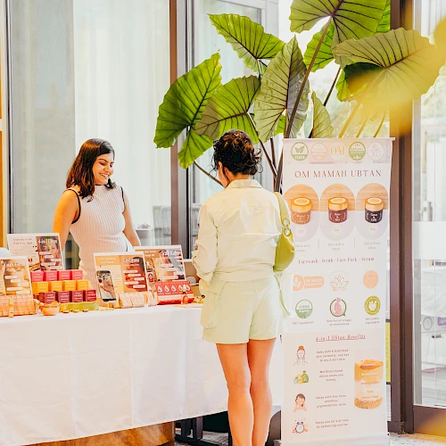 A woman stands at a booth with skincare products, chatting with a vendor inside a bright indoor market setup.