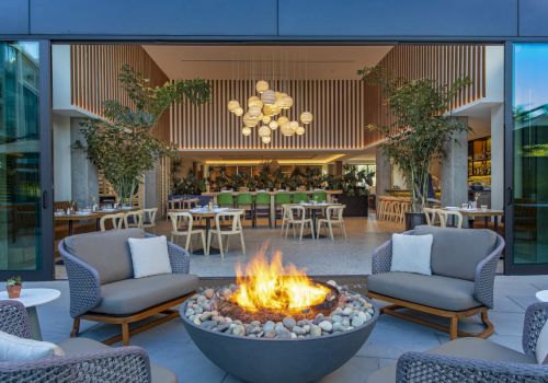 A modern outdoor lounge with a central fire pit, gray wicker seating, and a bright indoor dining area visible in the background, inviting and stylish.