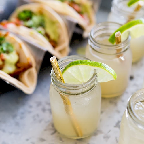 Margaritas with lime wedges in jars and tacos al pastor in the background.