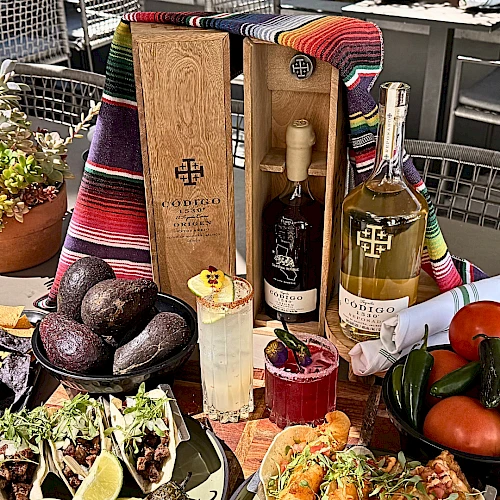 A colorful spread with two plates of tacos and avocado, tangy sauces, grilled corn, drinks (margarita and wine), and avocados plus tomatoes on a table.