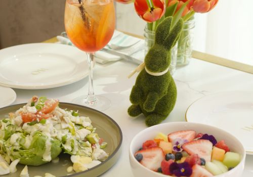 A bright table with brunch items: fruit-topped pancakes, a fruit salad, avocado-tomato salad, a bowl of berries, a fresh juice, and a vase of orange tulips shaped like a bunny, set near a window.