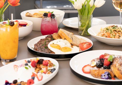 A sunny outdoor brunch table with eggs, pancakes, fruit, toast, yogurt, pastries, and colorful drinks, set beside flowers and wine glasses.