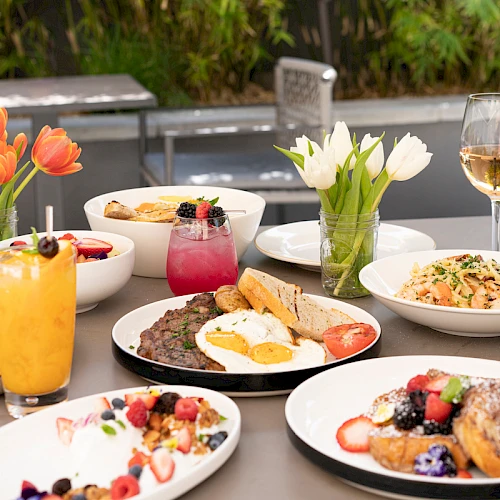 A sunny outdoor brunch table with eggs, pancakes, fruit, toast, yogurt, pastries, and colorful drinks, set beside flowers and wine glasses.