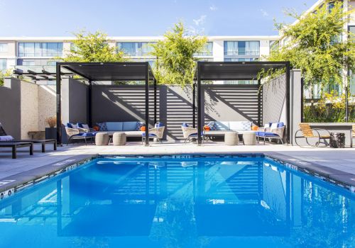 A modern poolside area with a bright blue rectangular pool, shaded loungers, and cabanas along the deck under blue skies.