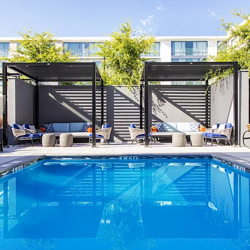 A modern poolside area with a bright blue rectangular pool, shaded loungers, and cabanas along the deck under blue skies.
