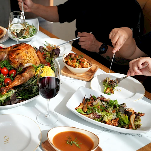 A family-style feast: roasted turkey, fresh salad, bowls of colorful sides, wine glass, and people sharing a plated meal at the table.