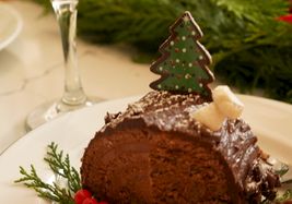 A festive chocolate cake slice on a plate with red berries and greenery, a Christmas tree decoration, and a cocktail glass in the background.