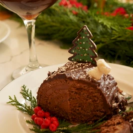A festive chocolate cake slice on a plate with red berries and greenery, a Christmas tree decoration, and a cocktail glass in the background.