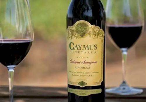 A bottle of Caymus wine on a table with two wine glasses, outdoors in sunlight, labeled Cabernet Sauvignon.