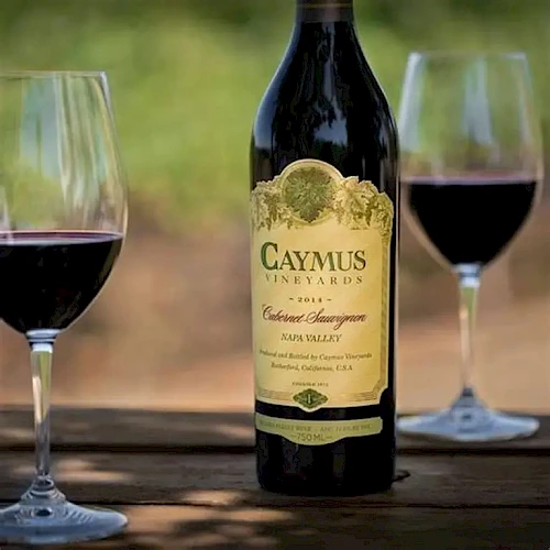 A bottle of Caymus wine on a table with two wine glasses, outdoors in sunlight, labeled Cabernet Sauvignon.