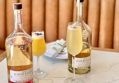 Two bottles of white wine with stems and two filled wine glasses on a marble table, a light brown wooden background, and a small booklet.