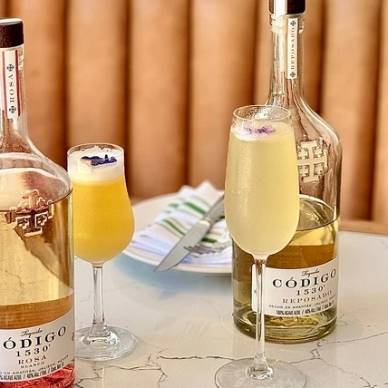 Two bottles of white wine with stems and two filled wine glasses on a marble table, a light brown wooden background, and a small booklet.