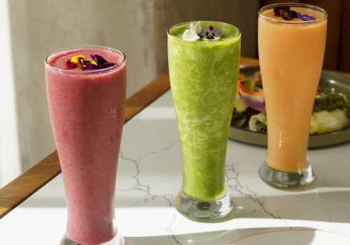 Three colorful smoothies in tall glasses on a marble table, pink, green, and orange, garnished with fruit, sunny casual vibe.